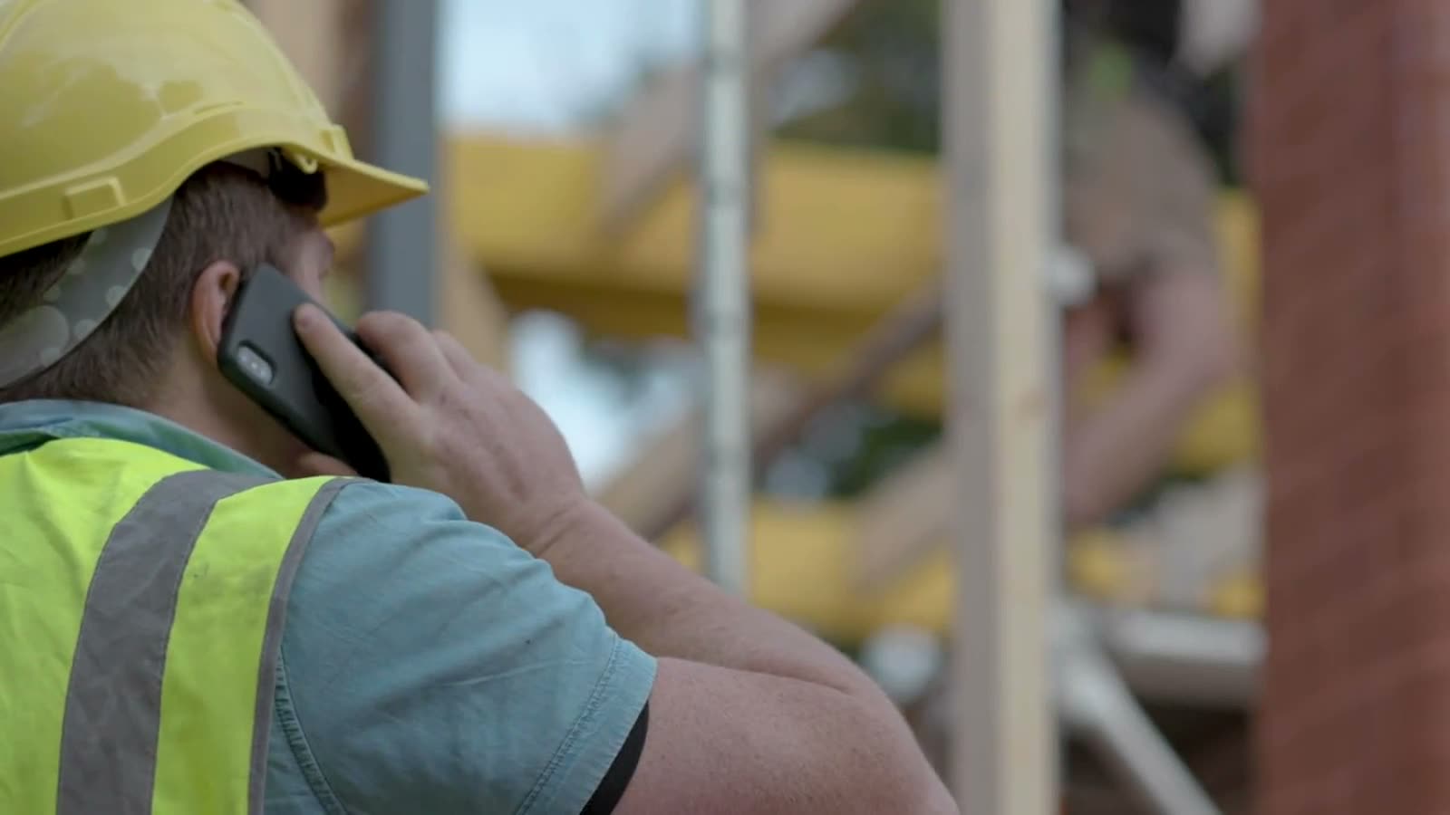 A construction worker wearing a hard hat and hi-vis vest uses a phone on site